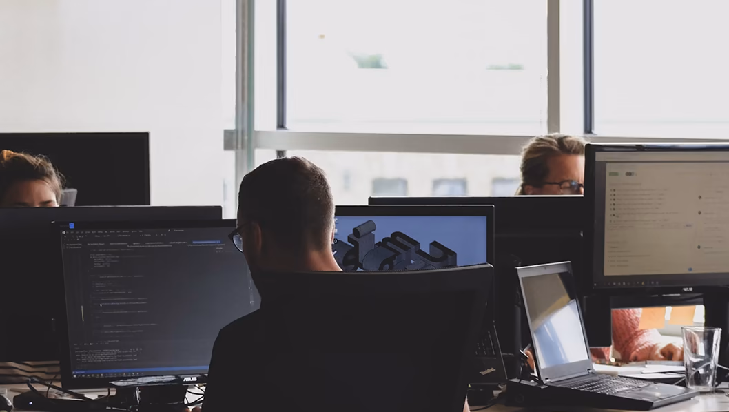An open-plan office of engineers at desks, daylight from a wall of windows behind them.